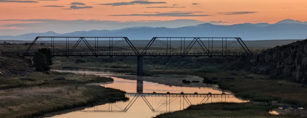 Restored historic steel truss bridge over a river, at sunset, with mountains in the background. Costilla Crossing Bridge over the Rio Grande (April, 2026 CGS Geology Calendar). Photo credit: Nate Rogers (CGS).