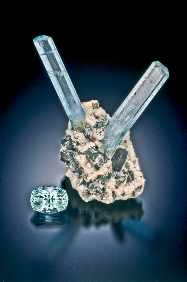 A typical specimen of aquamarine found on Mt. Antero, Teller County, with two elongate hexagonal crystals in a matrix of feldspar, mica, and smoky quartz, alongside a faceted stone. Photo credit: Jeffrey A. Scovil.